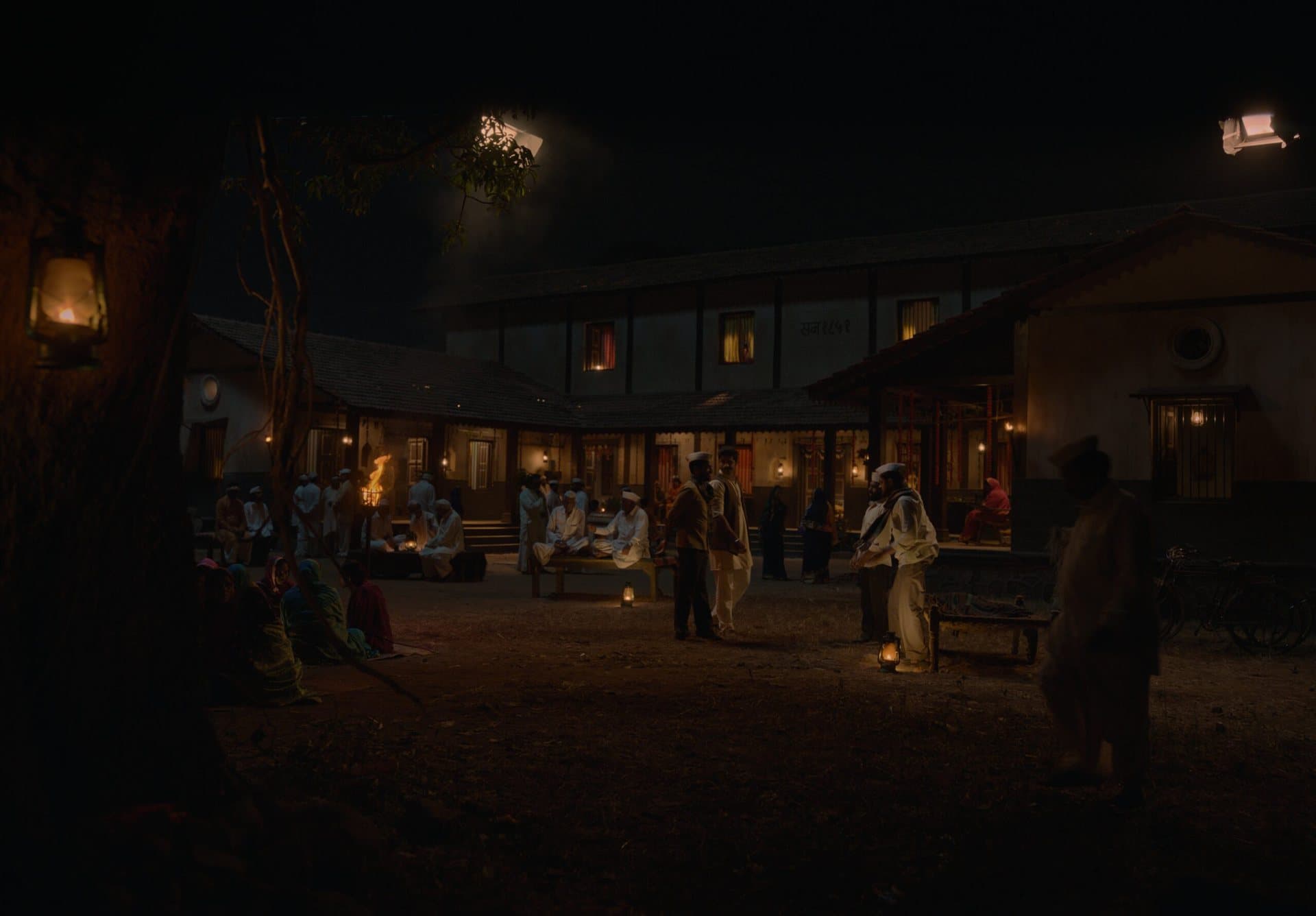 A two-storey village mansion with tiled roofs and lit windows at night; villagers gather in the dusty courtyard around lanterns and a small fire.