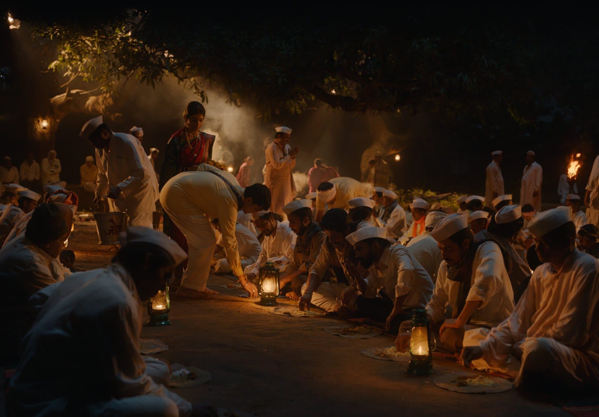 Men in white kurtas and Gandhi caps sit in rows eating from leaf plates by green kerosene lanterns, while a woman in a red sari serves under a large tree.