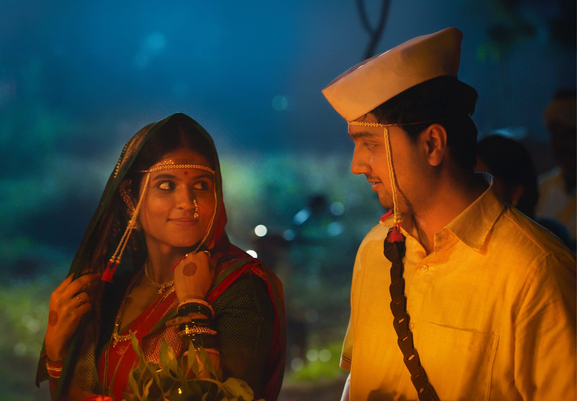 A bride in a red and green sari with pearl mundavalya looks sideways at a groom in a yellow shirt and white topi, lit by warm light against a blue dusk.